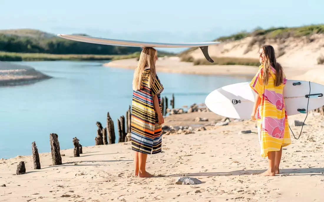 Für Surfer und Strandfreunde ist ein Surf Poncho quasi ein Muss Surferinnen im Surf Poncho und mit Surfbrett am Strand